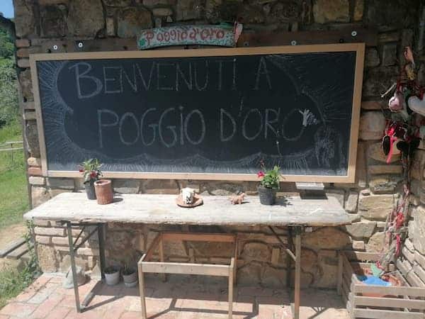 CEN - Poggiodoro - Lavagna in ardesia appesa al muro esterno di una casa in mattoni; la lavagna reca la scritta "Benvenuti a Poggiodoro". Sotto alla lavagna c'è un vecchio tavolo da sagra con una panca.